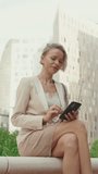 VERTICAL VIDEO, Businesswoman with blond hair wearing beige suit using mobile cellphone sitting outside on the street