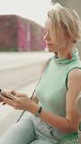 VERTICAL VIDEO, Beautiful woman with short blond hair in casual clothes sits at public transport stop, looks around, uses mobile phone