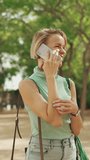 VERTICAL VIDEO, Beautiful smiling woman with short blond hair in casual clothes walks through the city square, talking on cellphone