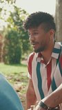 VERTICAL VIDEO: Close-up of young man with curly hair wearing striped shirt sitting in park having picnic on summer day outdoors, talking with friends