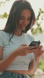 VERTICAL VIDEO: Young beautiful smiling woman with long brown hair wearing white crop top sitting in park having picnic on summer day outdoors, talking with friends, using cellphone