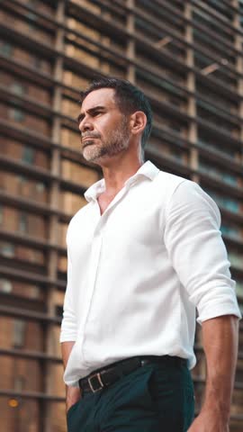 Mature businessman with neat beard wearing white shirt on his way to the office in the financial district in the city. Successful man Looks at the upper floors of modern buildings