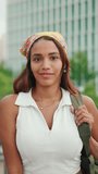VERTICAL VIDEO: Cute tanned woman with long brown hair wearing white top and yellow bandana posing while standing on bridge. Smiling girl looking at the camera