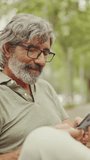 VERTICAL VIDEO: Middle-aged man with gray hair and beard wearing casual clothes sits on bench and uses mobile phone. Mature man in eyeglasses raises his head, looks at the camera and smiles