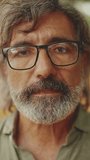VERTICAL VIDEO: Clouse-up, friendly middle-aged man with gray hair and beard wearing casual clothes looks at the camera. Mature gentleman in eyeglasses smiles at the camera