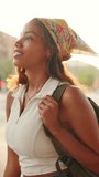 VERTICAL VIDEO: Cute tanned woman wearing white top and yellow bandana with backpack on her shoulder walks down the street against modern building background. Backlight