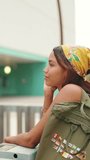 VERTICAL VIDEO: Profile of cute tanned woman with long brown hair wearing white top and yellow bandana stands on bridge with backpack on her shoulder and looks at modern city