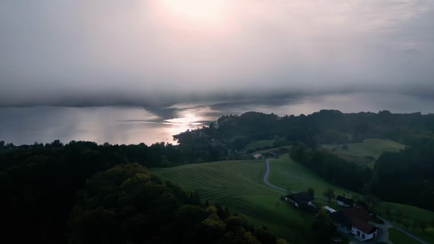 An aerial view of a small, rectangular house with a red roof and a wooden dock, located on the edge of a calm, blue lake surrounded by lush green trees under a partly cloudy sky