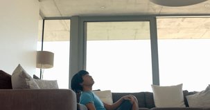 Teenage Asian boy sits comfortably on a couch at home, with copy space. He is focused on a digital tablet, engaged in online learning or leisure, slow motion.