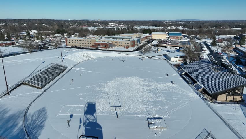 Aerial shot of a high school sports field covered in snow. Football stadium and track with bleacher seating. Winter in America theme.