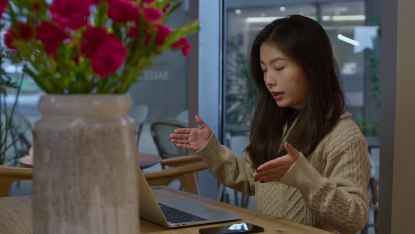 Young pretty Asian chinese girl having online call, gestures with hands. Work remotely from cafe.