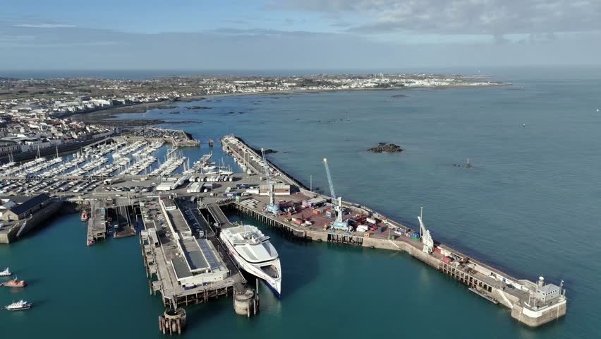 St Peter Port Guernsey flight over ferry terminal with ferry in dock, commercial dock with cranes towards Salerie Corner and over QE II Marina with views over Belle Greve Bay on bright sunny day Royalty-Free Stock Footage #3450518119