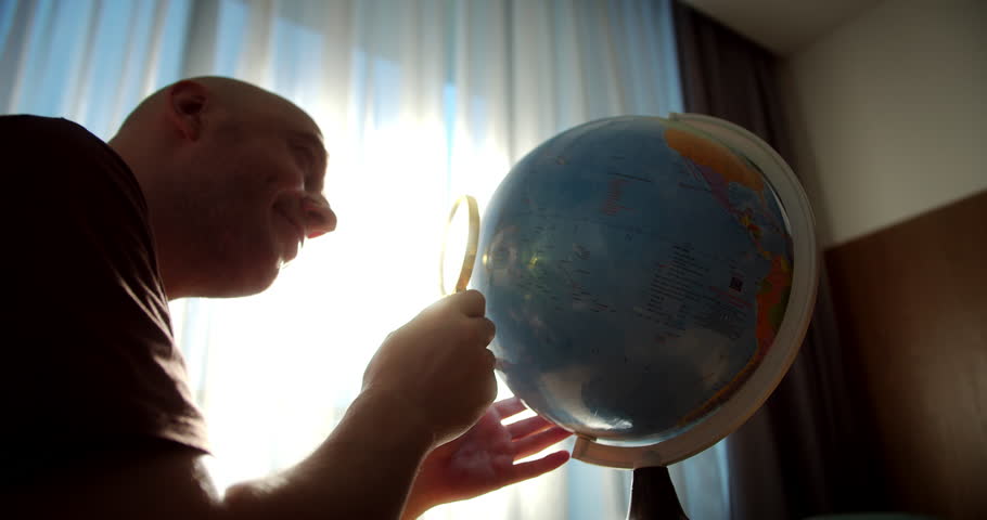 Man looks through a magnifying glass at a globe. Man using globe sits at home looking for right country on globe. 4K