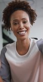 Vertical shot of a biracial woman in an office having a video call, smiling and wearing earbuds. Corporate environment concept.