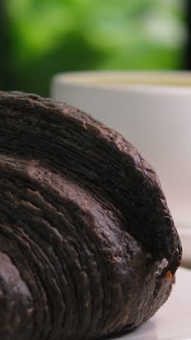 black croissant with cuttlefish ink on a white plate with a cup of cappuccino latte coffee against a green window background summer breakfast, France gourmet food delicious lunch snack