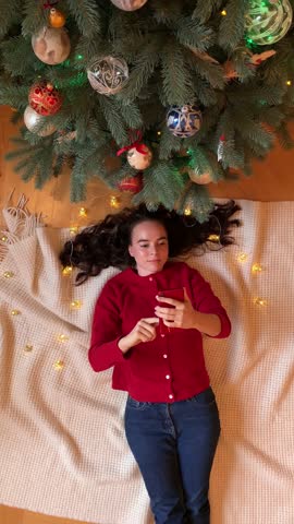 Top view of a beautiful young woman lying near Christmas tree, smiling, holding and scrolling her phone.