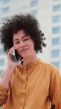 Vertical individual close up portrait of a young latin woman with curly hair having a call by smart phone. Hispanic lady with yellow shirt talking by cellphone sitting outdoors. Communication concept