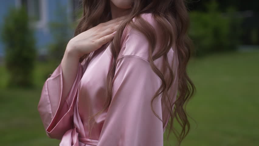 Woman in a pink robe standing in grass with layered hair and a formal gesture