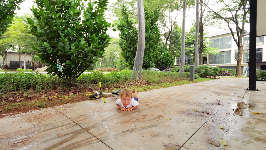 Cheeky toddler lies down on ground after runbike slip, dramatically pretending to be injured. With exaggerated and mischievous expression, he awaits sympathy for minor incident. Camera move closer. Royalty-Free Stock Footage #3450935541