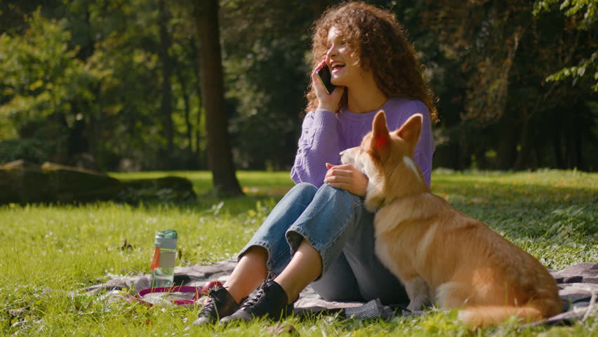 Happy Caucasian young girl pet friendly woman female handler joyful lady talking mobile phone laughing playing with cute lovely dog welsh corgi puppy in city park green forest summer nature outdoors Royalty-Free Stock Footage #3451069353