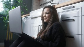 Young asian woman sitting on the floor in kitchen having video call conversation through using laptop computer. Share news, talk to family, enjoy webchat virtual meeting