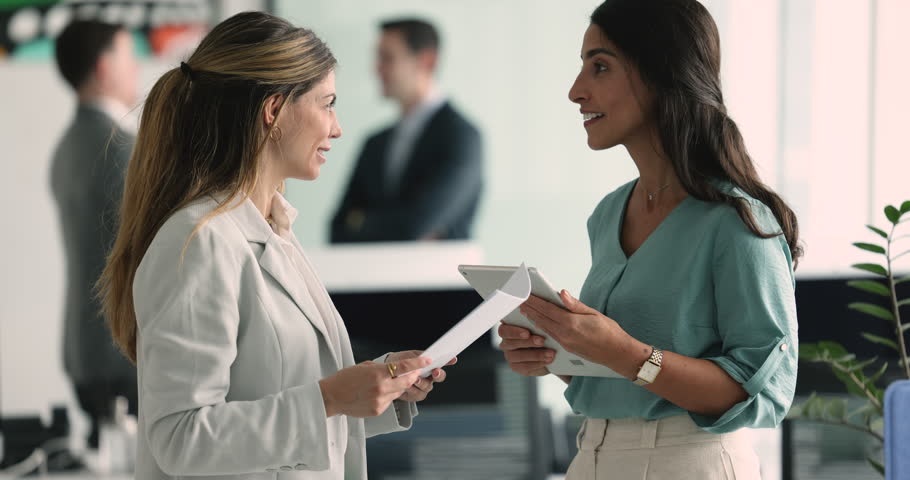 Two young attractive businesswomen colleagues discuss contract details, smile, enjoy conversation walk along hallway in modern office, share ideas, opinion, solutions, engaged in teamwork and workflow