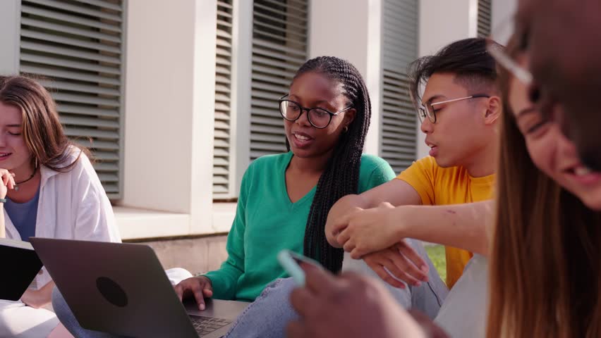 Group of young international university students using laptop on the grass outside the faculty building. Friends gathered happily studying together on a park at campus