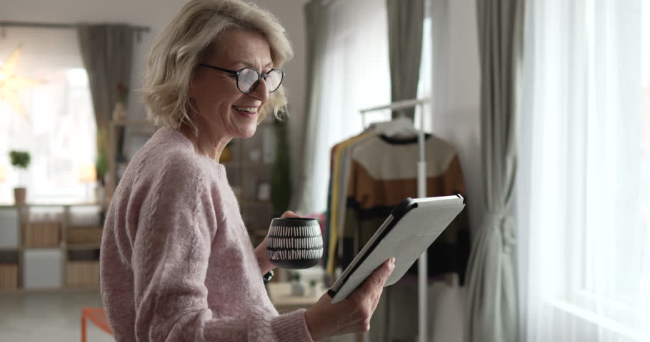Portrait of mature blonde caucasian woman with eyeglasses use digital tablet at home browse internet or watch online video happy smile slow motion video