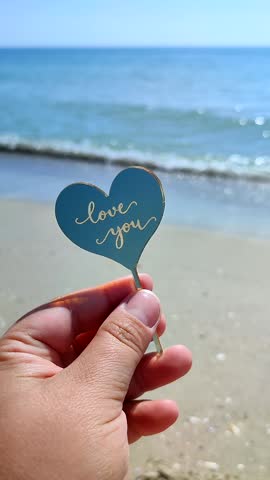 Person holding in fingers plastic stick in shape heart and words Love you on background sand beach and sea waves in seashore on sunny summer day. Concept love romance amour St Valentines Day Vertical