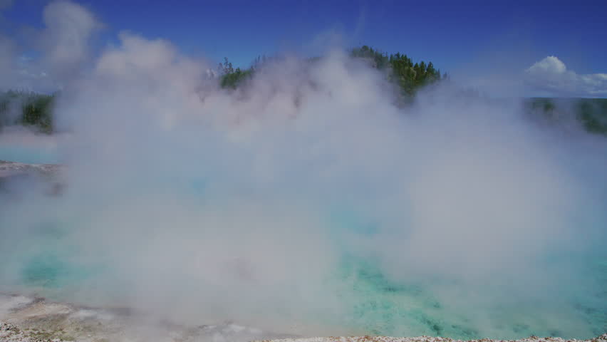 Thick clouds of steam envelop the geothermal features of Yellowstone National Park, with visitors barely visible on the boardwalk under a bright blue sky. Slow Motion, 4K RAW. 