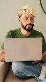 Vertical video, Young blond man with a beard wearing an olive green T-shirt works on a laptop while sitting in a cozy living room