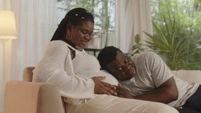 Medium shot of black happy husband listening to baby quickening in belly of pregnant wife sitting on couch - Powered by Shutterstock - Get 15% off with code: PIKWIZARD15