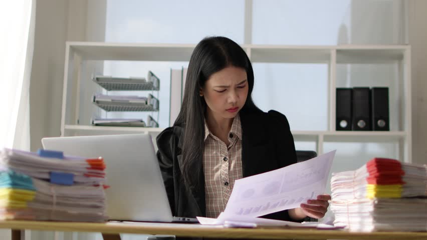 Serious Asian businesswoman is busy with paperwork on a messy desk. Woman are stressed due to excessive workload and are forced to overworked. Royalty-Free Stock Footage #3451648973