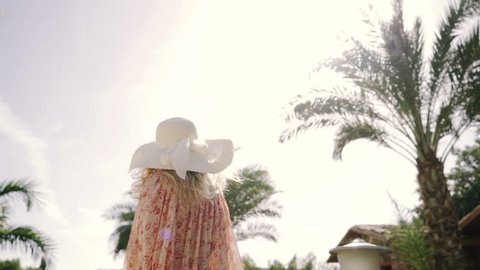 Summer lifestyle, pretty young woman walking near resort on the tropical island with palm. Wearing stylish shirt dress, sunglasses, straw hat. Travel and journey