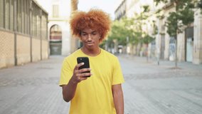 Young African-American man using mobile phone while walking smiling down city street. Slow motion video of smiling young afro man writing messages and surfing the internet with his smartphone 