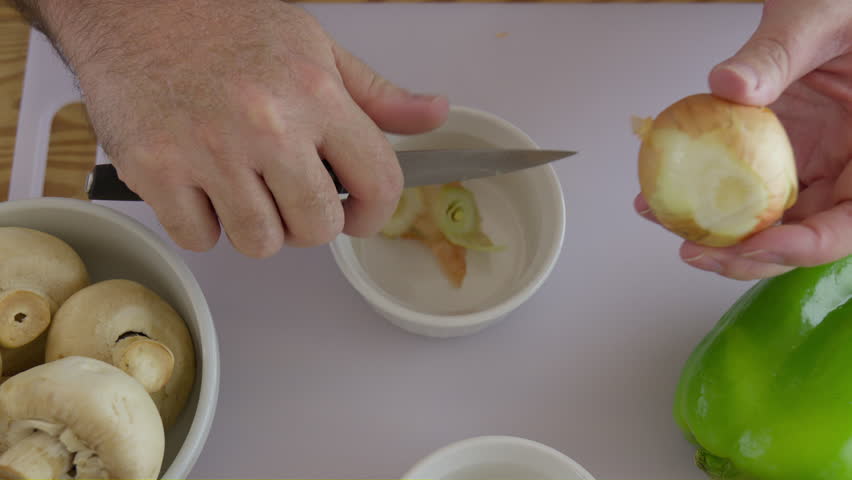 Peeling onion skin with knife and hand on white cutting board with green bell pepper and mushroom at side