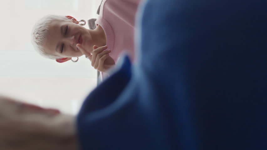 Vertical over shoulder of two young Caucasian women sitting in front of each other in apartment and having conversation with help of sign language