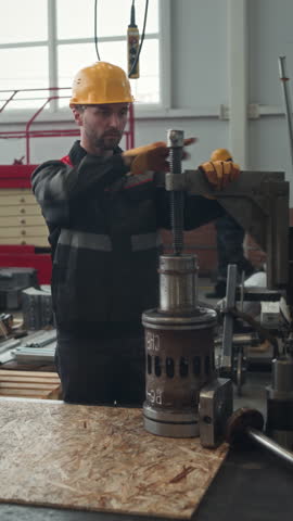 Vertical medium shot of Caucasian male worker in workwear and yellow hardhat repairing machine equipment at industrial plant