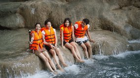 Young Asian man and woman friends enjoy and fun outdoor lifestyle travel nature forest using action camera taking selfie vlogging during playing water at waterfall lagoon on summer holiday vacation.