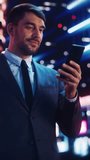 Vertical Screen: Portrait of a Handsome Man in Stylish Suit Standing in a Modern City Street with Neon Lights at Night. Attractive Male Using Smartphone and Looking Around the Cinematic Environment.