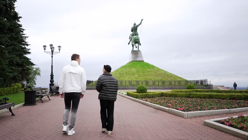 A man and a woman in jackets walk through the park, with a monument in the background.