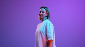 Beautiful young girl in white t-shirt standing, posing with smile against purple studio background in neon light. Video, 4k. Concept of youth, human emotions, casual fashion, lifestyle
