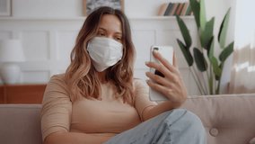 alone attractive native American young woman with medical mask sits on sofa at home, holding mobile phone in hands and making selfie photo looking in front camera