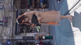 Vertical video. Asian young woman business lady with a folder for documents walks down the street in a raincoat with a fashionable silk scarf around her neck against the backdrop of a business center 