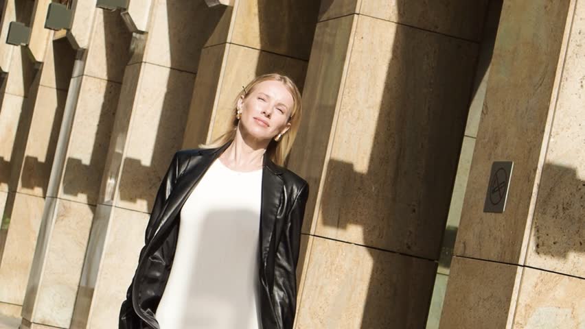 Beautiful fashionable blonde woman walking on street in city, wears leather jacket, hard sunlight