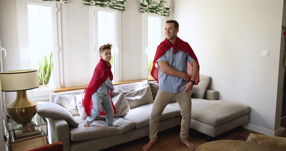 Carefree daddy and son dance, play together at home. Little boy and dad wear red cloaks pretend superheroes, having funny playtime, move to pop music, jumping, fool around in living room, full-length Royalty-Free Stock Footage #3452257511