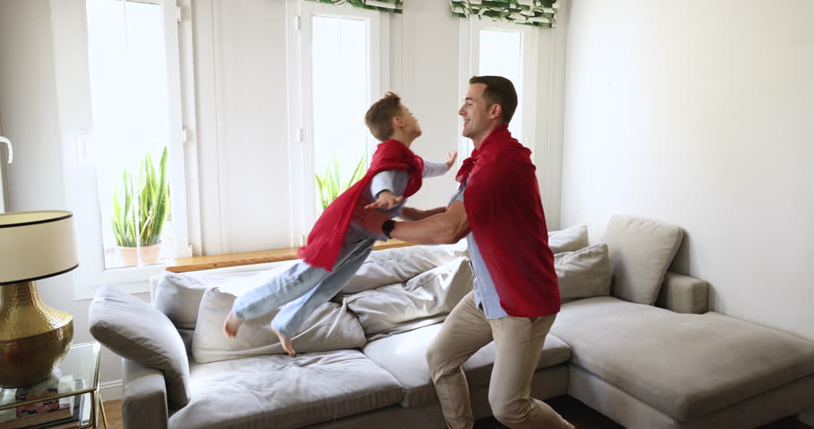 Happy young father playing with little son at home, wear red cloaks looks like superheroes having fun together in modern cozy living room. Cute boy jumping, enjoy playtime with loving dad on weekend Royalty-Free Stock Footage #3452269001