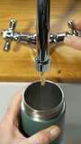 Vertical video – Cinemagraph - Video of fresh running water flow from a sink tap or faucet, combined with a still image of a hand holding an insulated steel bottle as it fills up.