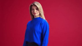 Beautiful young woman with blonde hair, wearing blue sweater, looking at camera and smiling against red studio background. Happiness. Concept of human emotions, youth, fashion, expression