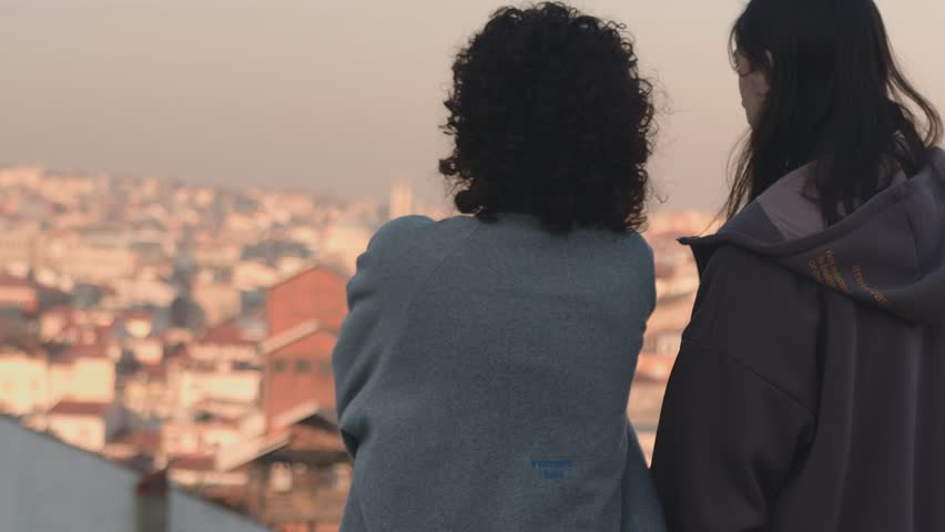 This frame captures a tender moment between two individuals set against the soft, early morning light that bathes the cityscape in the background. The focus is on the two friends, one with curly hair Royalty-Free Stock Footage #3452345525
